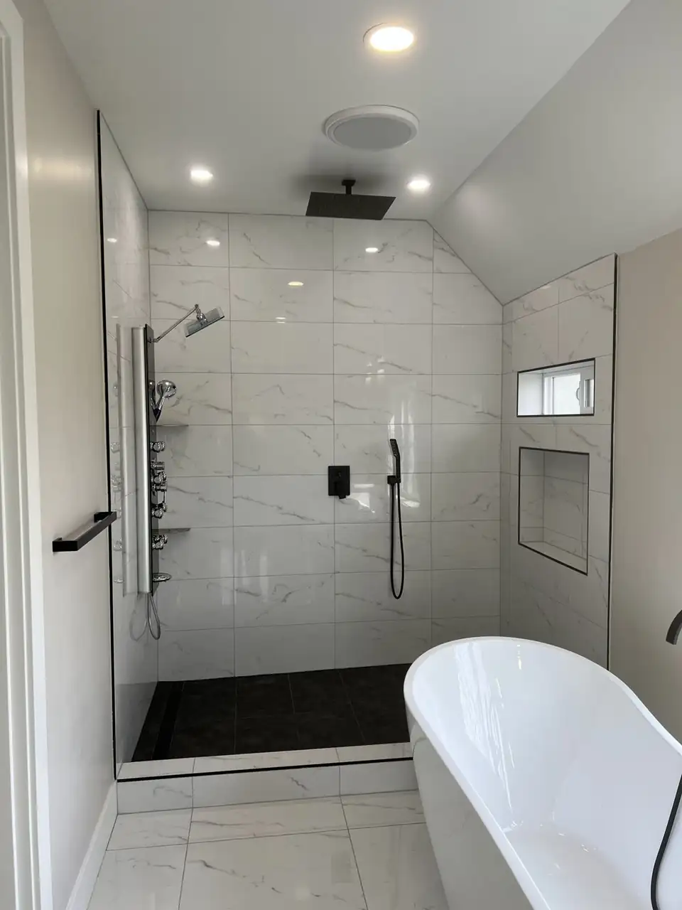 Walk-in shower by Model Tile, bathroom remodeler in Everett, WA: white marble-look porcelain walls, black floor tile with a linear drain, matte black fixtures and a freestanding tub